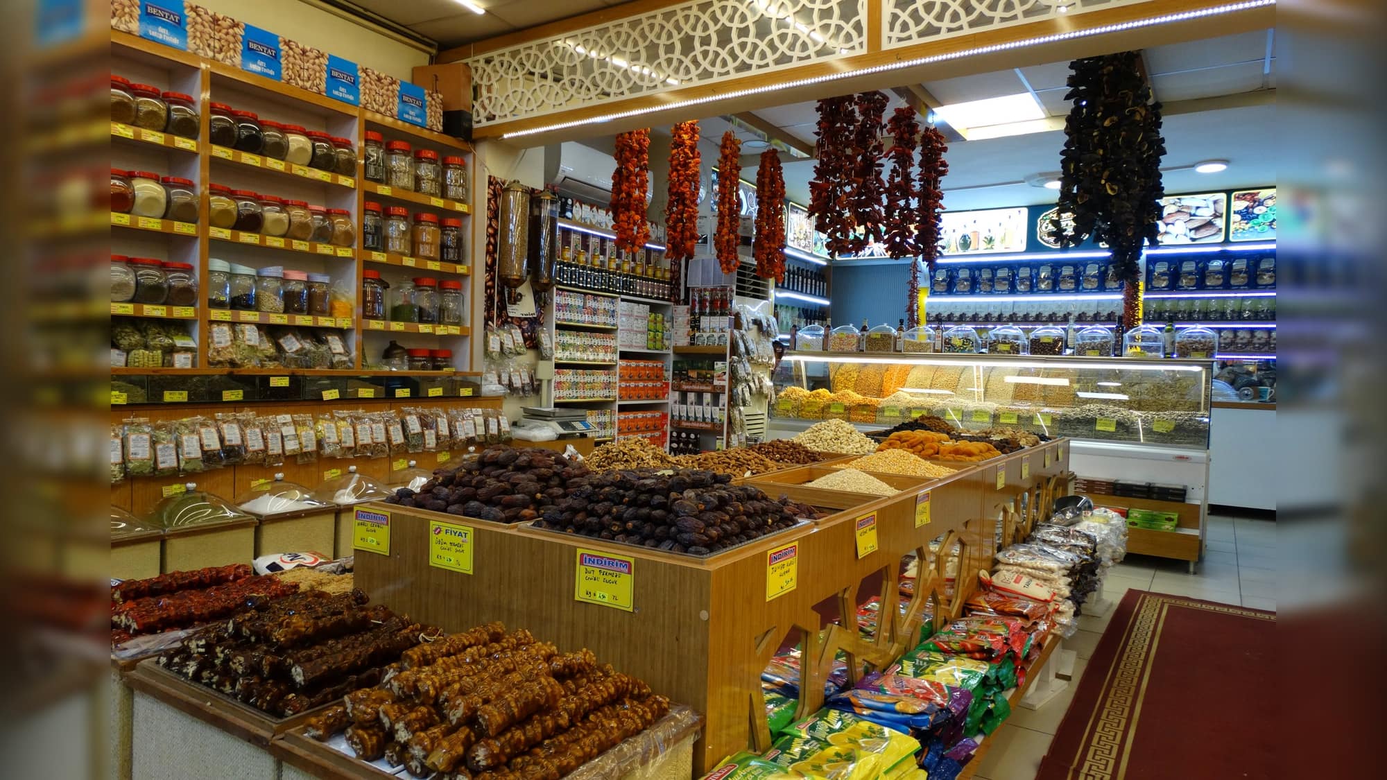 Dükkanın içi: baharat rafları, tavanda kurutulmuş biber dizisi ve tezgahta kuru meyve yığınları